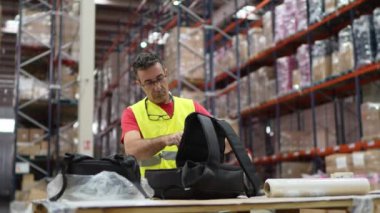 Çelik yelek giyen bir depo işçisi yoğun bir depo ortamında nakliye için dikkatlice bir paket hazırlıyor.