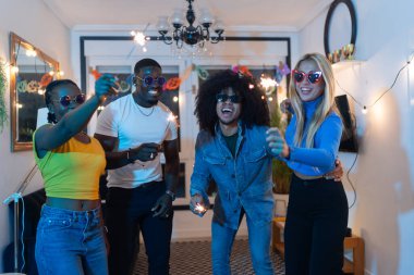 Çok ırklı arkadaşlar, neşeli bir ev partisinden zevk alıyorlar, dans ediyorlar ve neşeli ifadeli maytap sallıyorlar.