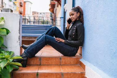 Genç Latin kadın basamaklarda oturuyor, telefonla konuşuyor ve şehir ortamında bir anlık rahatlığın tadını çıkarıyor.