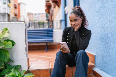 Genç Latin Amerikalı kadın bir terasın merdivenlerinde otururken cep telefonu kullanıyor.