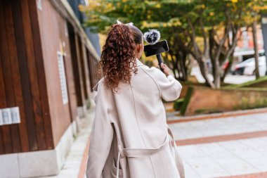 Genç Latin kadın vlogger yürüyor ve akıllı telefon ve mikrofonla şehir caddesinde yürüyor ve çekim yapıyor.