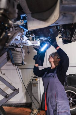 Otomobil tamirhanesinde asansörle kaldırılan aracın altını incelerken el feneri kullanan Çinli kadın tamirci.