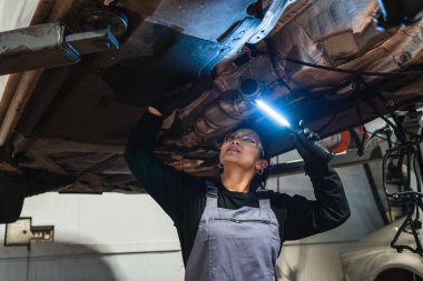 El feneri kullanan kadın tamirci, oto tamirhanesinde asansörle kaldırılan aracın altını inceliyor.