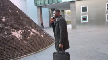 Profesyonel afrikalı işadamı telefon görüşmelerini yaparken güvenli bir şekilde şehir manzarasında geziniyor, modern kurumsal başarıyı ve dinamik profesyonel yörüngeyi somutlaştırıyor.