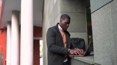 Genç Afrikalı girişimci mobil teknolojiyi kullanıyor, açık havada dizüstü bilgisayarda çalışıyor ve şehir profesyonel ortamında dinamik yavaş çekim etkisi yaratıyor.