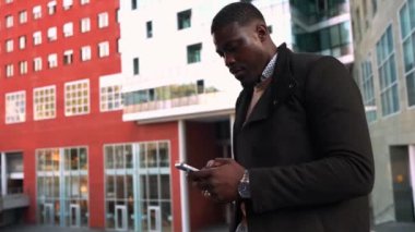 Profesyonel Afrikalı iş adamı modern şehir gökdelenleri yakınında akıllı telefon kullanıyor, dijital girişimciliği somutlaştırıyor ve kentsel başarıyı yavaş çekimde yaşıyor.