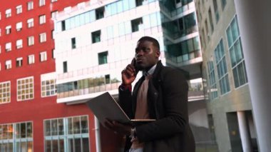 Genç Afrikalı girişimci dizüstü bilgisayar kullanıyor ve modern ofis binalarının yakınında telefonla konuşuyor.