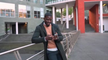 Genç profesyonel afrikalı iş adamı, modern ofis binasının yakınındaki akıllı telefona bakıyor, parmaklıklara yaslanıyor ve ağır çekimde gülümsüyor.