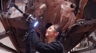 Profesyonel kadın tamirci oto tamirhanesinde el feneri kullanarak arabanın bagajını dikkatlice inceliyor.