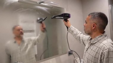Saç kurutma makinesi kullanarak banyo aynasındaki erkek yoğunlaşması sabah rutin sorununu yavaş çekimde kurutma tekniğiyle çözüyor.