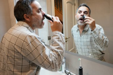 Orta yaşlı bir adam sabah rutini sırasında banyo aynasına bakarken burun kıllarını kesmek için küçük bir elektrikli makina kullanıyor.