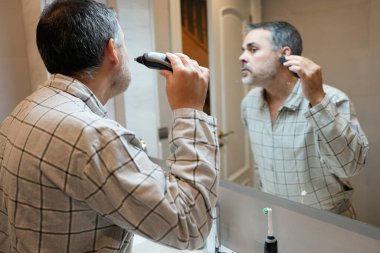 Olgun bir adam sabah banyodaki aynaya bakarken sakalını kesmek için tıraş bıçağı kullanıyor.