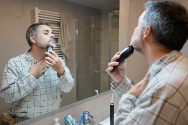 Pijamalı olgun bir adam, sabahları banyo aynasına bakarken sakalını tıraş etmek için elektrikli tıraş bıçağı kullanıyor.
