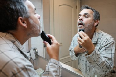 Yaşlı adam sabah banyodaki aynaya bakarken sakal tıraşı için jilet kullanıyor.