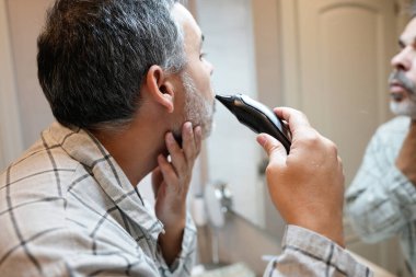 Yaşlı adam sakalını kesmek için elektrikli tıraş makinesi kullanıyor, tuvaletteki görünüşüne dikkat ediyor.