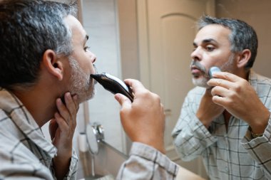 Orta yaşlı bir adam sakalını elektrikli jiletle kesiyor. Sabah rutini sırasında banyo aynasına yansıyor.