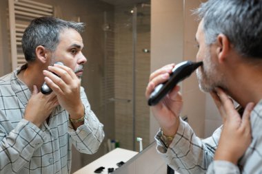 Kıdemli adam sakalını kesmek için elektrikli tıraş makinesi kullanıyor. Sabah rutini sırasında banyo aynasına yansıyor.