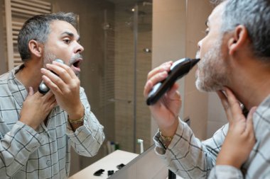 Olgun bir adam, banyo aynasının önünde sakalını jiletle keserken şaşırmış bir ifade takınıyor.
