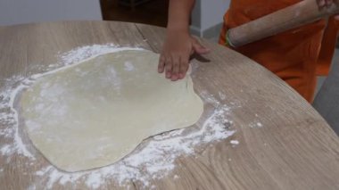 Çocuk turuncu önlük giyip pizza hamurunu yassılaştırıyor ve ev mutfağındaki ahşap masa üzerinde merdane takıyor.