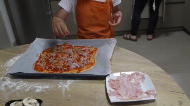 Neşeli çocuk, ev yapımı pizza hazırlıyor. Ev mutfağında aşçılık becerilerini öğrenirken, taze hamurun üzerine dikkatle jambon katlıyor.
