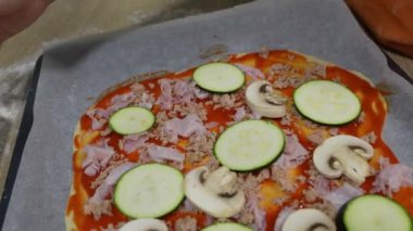 Genç aile üyeleri domates sosunu yayarken, kabak, mantar ve jambonu ev mutfağındaki taze pizza hamuruna eklerken iş birliği yapıyorlar.