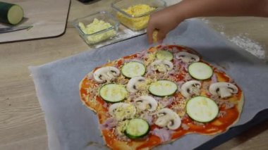 Genç aşçı ev mutfağındaki pizza tabanına dikkatlice kabak, mantar ve peynir ekleyerek ev yapımı pizza hazırlıyor.