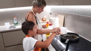 Annesi, genç oğlunun da yardımıyla, modern bir mutfakta, kızgın tavaya mısır tanelerini dikkatlice dolduruyor.