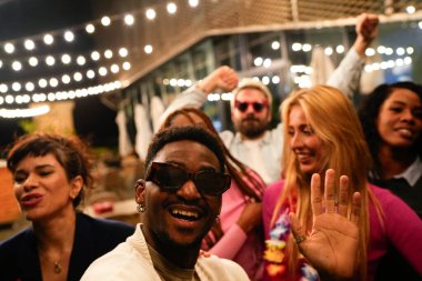 Genç çoklu etnik arkadaşlar canlı bir çatı partisinde neşeli bir şekilde dans ediyorlar. Etrafı ışıldayan telli ışıklar ve şenlikli bir atmosferle çevrili.