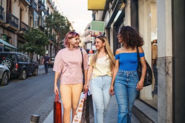 Alışveriş torbaları taşıyan ve alışveriş çılgınlığından sonra sokakta yürüyen üç mutlu bayan arkadaş.
