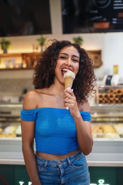 Kıvırcık saçlı bir kadın dondurmacıda leziz bir dondurma külahının tadını çıkarıyor, tatlı ikramının tadını çıkarırken neşeyle gülümsüyor.
