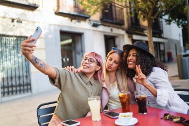 Üç neşeli bayan arkadaş, açık bir kafede içecek ve atıştırmalıkların tadını çıkarırken akıllı telefonlarıyla selfie çekiyorlar.