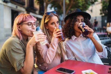 Üç genç kadın bir masada oturmuş, içki içiyor ve neşeli, açık havada sosyalleşiyorlar.