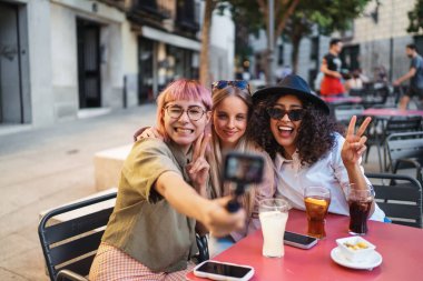 Three cheerful female friends are making peace signs while taking a selfie with a gimbal at a vibrant outdoor cafe clipart