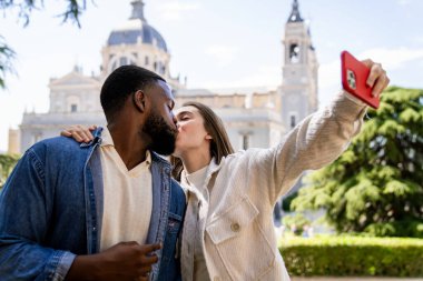 Şehirdeki bir katedralin önünde öpüşen ve akıllı telefonuyla selfie çeken çok ırklı bir çift.