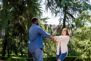 Neşeli, çoklu etnik çeşitlilikte bir çift el ele tutuşup, etrafında dönüyor, yeşil bir parkta güneşli bir günün tadını çıkarıyorlar.