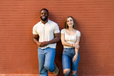 Mutlu çoklu etnik çift gülümseyen ve canlı kırmızı tuğla bir duvara yaslanan, günlük bir şehir ortamında neşe ve beraberlik saçan