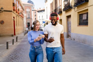 Çok ırklı genç çift akıllı telefon kullanıyor ve şehir caddesinde yürürken kulaklık takıyor.