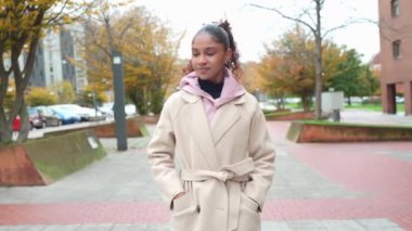 Genç çağdaş kadın şehir manzarasında geziniyor, akıllı telefon kullanarak güvenli bir şekilde yürüyor, modern dijital yaşam tarzını şık ve rahat hareketlerle somutlaştırıyor.