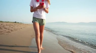 Genç çıplak ayaklı genç kumlu sahilde yürüyor, müzik dinliyor ve açık mavi gökyüzünün altında okyanus dalgalarıyla akıllı telefonlara bakıyor.