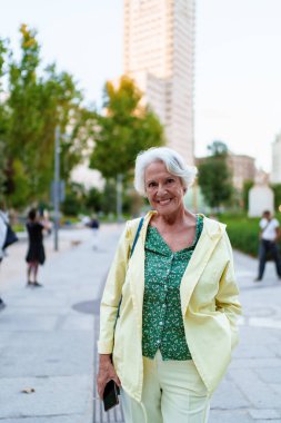 Elinde akıllı bir telefonla modern bir şehir parkında yürüyen, emekliliğin ve şehir hayatının tadını çıkaran şık bir son sınıf öğrencisinin portresi.