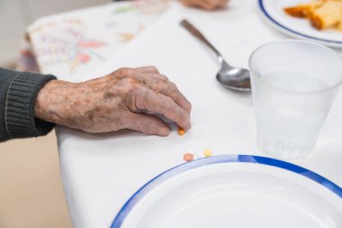 Huzurevi masasında haplar düzenleyen, günlük sağlık hizmetleri rutinlerini ve kamu ortamında üst düzey bakım yönetimini vurgulayan yaşlı kişi