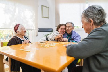 Huzurevindeki yaşlı kadınlar, sıcak, güneşli bir odada refakatçilik ve neşe besleyerek, hareketli bir sohbet ve oyunla meşgul olurlar.