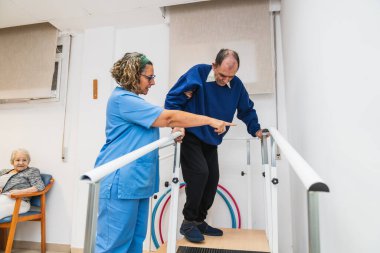Hemşire, huzurevindeki paralel barlarda, yaşlılara rehabilitasyon ve yaşlılara destek olmak için hareket egzersizleri yapan yaşlı bir adama yardım ediyor.
