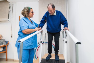 Hemşire, huzurevindeki paralel barlarda yapılacak rehabilitasyon seansında yaşlı bir adamı destekliyor, yaşlıların taşınabilirliğini ve bakımını teşvik ediyor.
