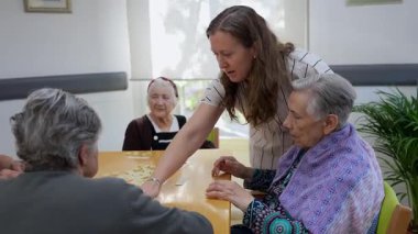 Huzurevinde fayanslarla kelime oyunu oynayan yaşlı kadınlara yardım eden yaşlı hemşire, bilişsel becerileri ve sosyal etkileşimi teşvik ediyor