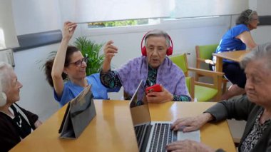 Yaşlılar ve bir bakıcı, huzur evinde müzik dinleyip dans ederek modern teknolojiyi kullanarak, neşeli bir anı paylaşıyorlar.