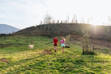 Gün batımında güzel yeşil bir çayırda köpekleriyle yürüyen genç çift, doğanın huzur ve sessizliğinin tadını çıkarıyorlar.