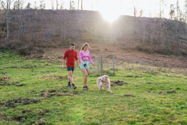 Sporcular, tüylü arkadaşlarıyla birlikte güneşin sıcacık parıltısıyla güneşlenen kırsal bir yerde rahatlatıcı bir yürüyüşün tadını çıkarıyorlar.