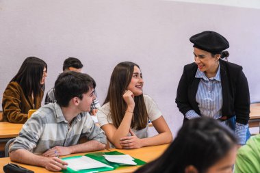 Gülümseyen bayan öğretmen bir ders sırasında lise öğrencilerine yardımcı oluyor, sınıf ortamında rehberlik ve destek sağlıyor.