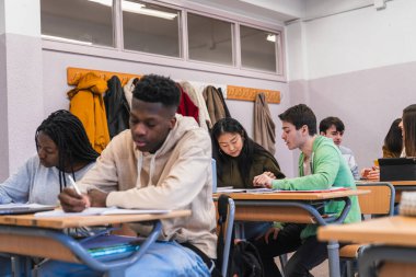 Çok ırklı lise öğrencileri birlikte çalışıp notlar alırken, aynı zamanda canlı bir sınıf ortamında öğrenme faaliyetlerinde bulunuyorlar.